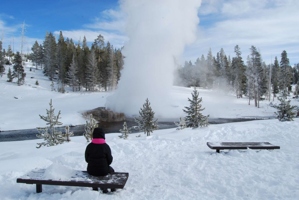 Woman sitting on bench watching Riverside Geyser erupt in winter