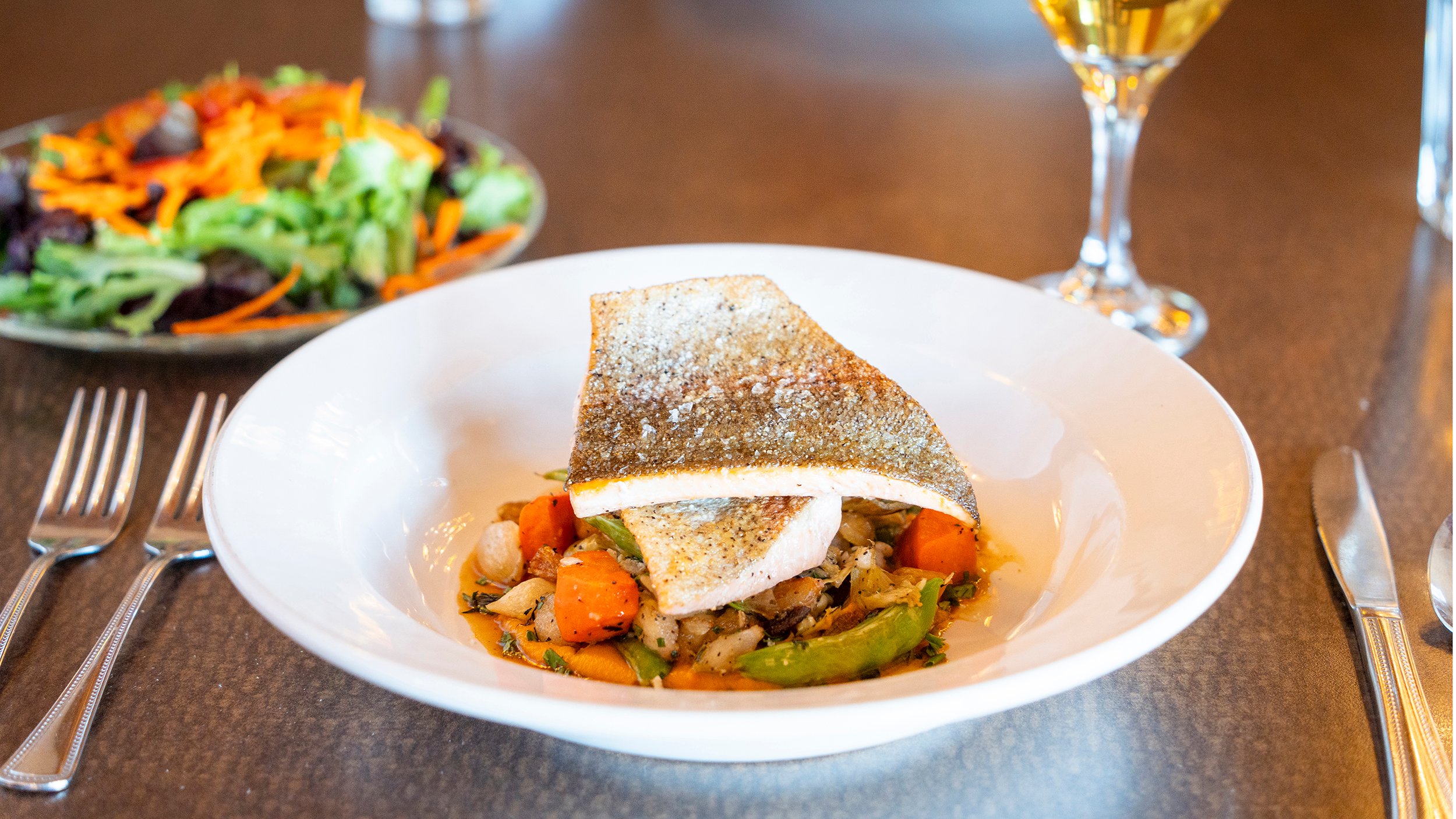 Table with cooked trout, salad, and glass of white wine